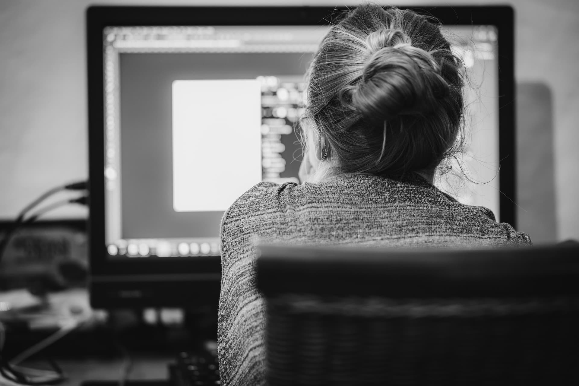 Black-and-white photo from behind a woman working at her desktop computer in a home office
