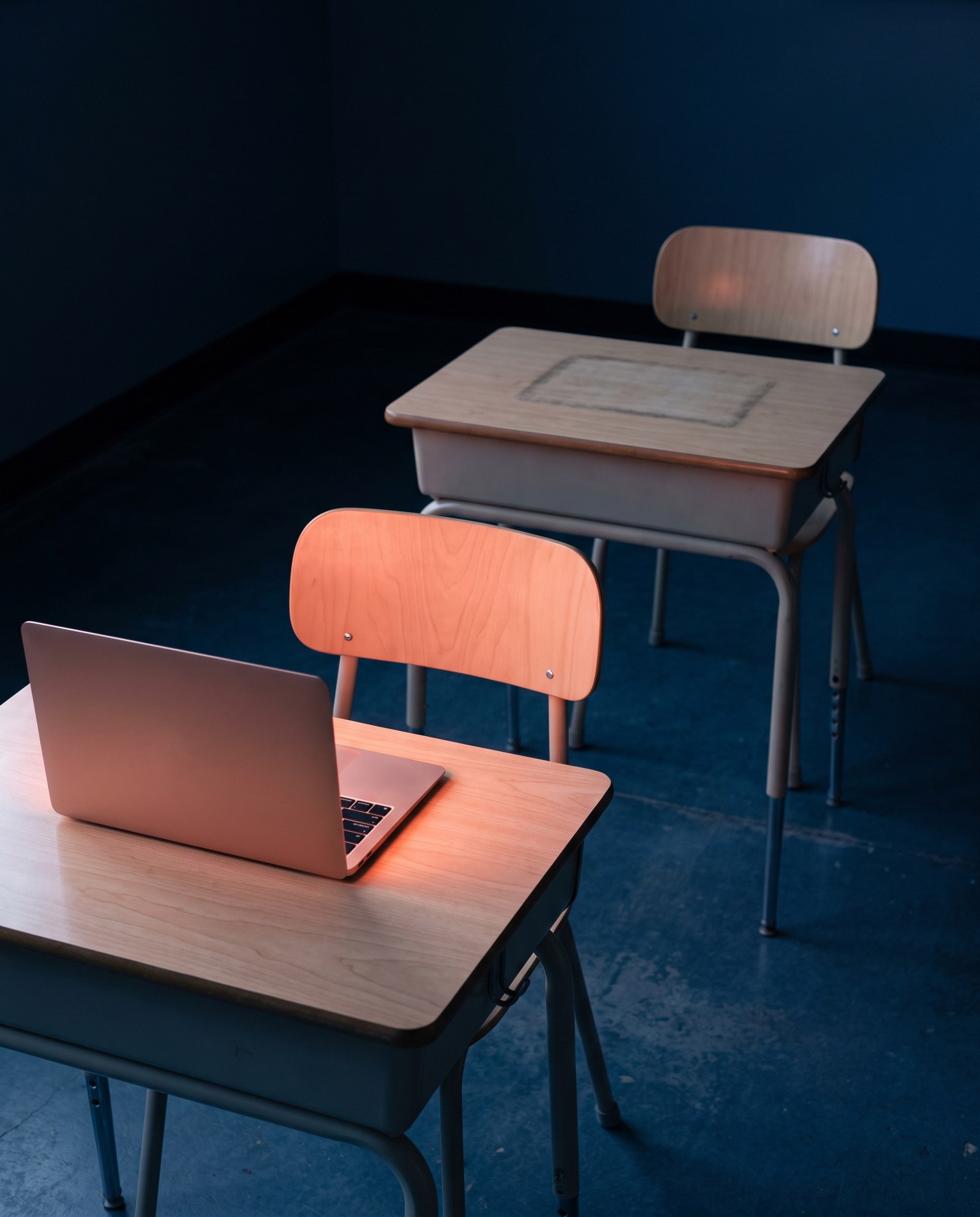 Two empty school desks in a dark room with deep navy walls. On the near desk, an open laptop with its screen turned away spills warm coral light across the wood. The far desk is bare except for a faint rectangular outline, the size of the laptop, worn into its surface.